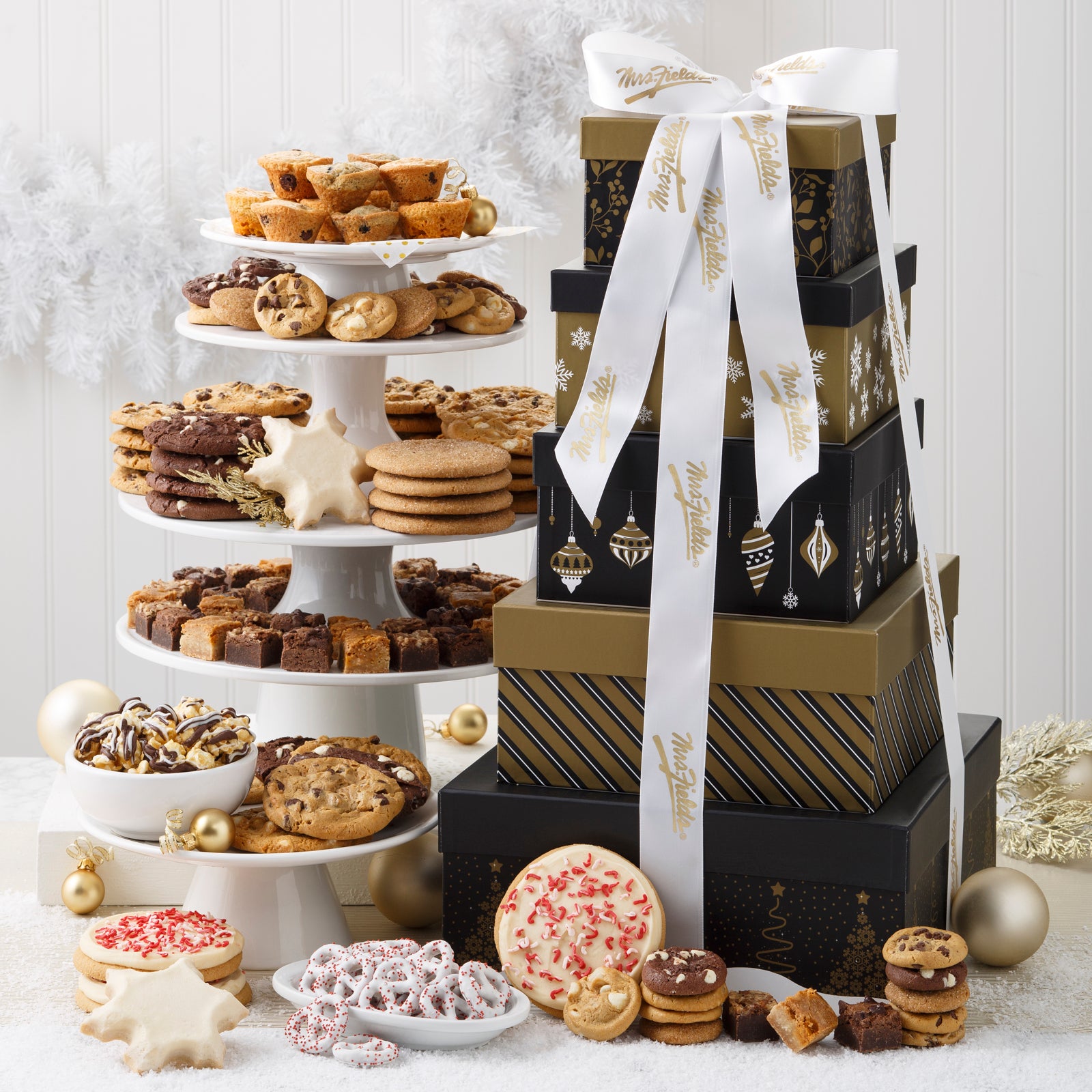 A five-tier gold and black square tower tied at the top with a white Mrs. Fields ribbon and surrounded by an assortment of Nibblers®, original cookies, brownie bites, muffins, three frosted peppermint cookies with sprinkles, three frosted snowflakes, yogurt-covered pretzels, and chocolate drizzled popcorn.