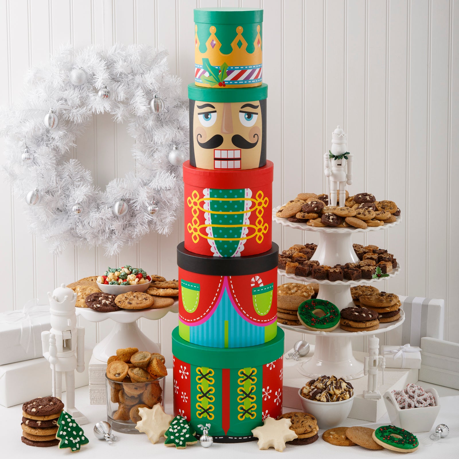 A five-tier nutcracker tower surrounded by an assortment of Nibblers®, original cookies, muffins, holiday popcorn, chocolate drizzled popcorn, yogurt-covered pretzels, two frosted wreath cookies with sprinkles, two snowflake cookies, and two frosted holiday trees with sprinkles.