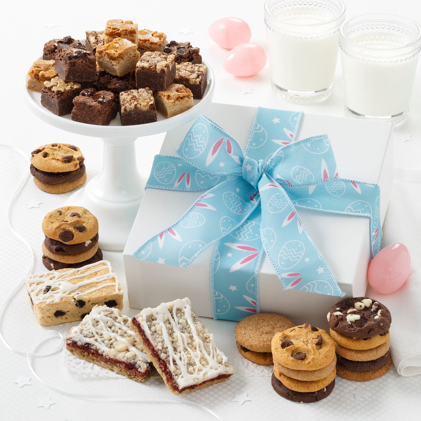 A white gift box tied with a light blue ribbon decorated with bunny ears and surrounded by an assortment of nibblers, brownie bites, and fruit bars