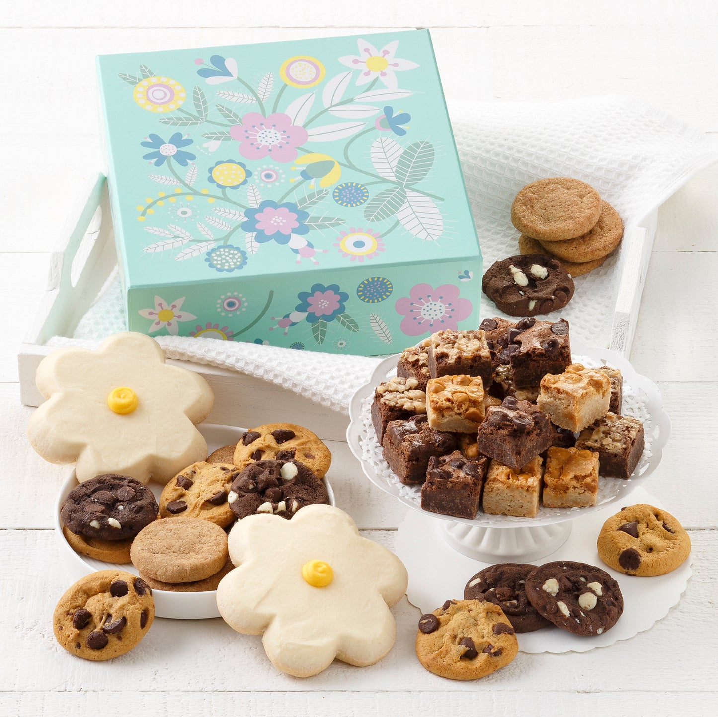 A light blue gift box decorated with flowers and surrounded by an assortment of nibblers, brownie bites, and two frosted daisy cookies