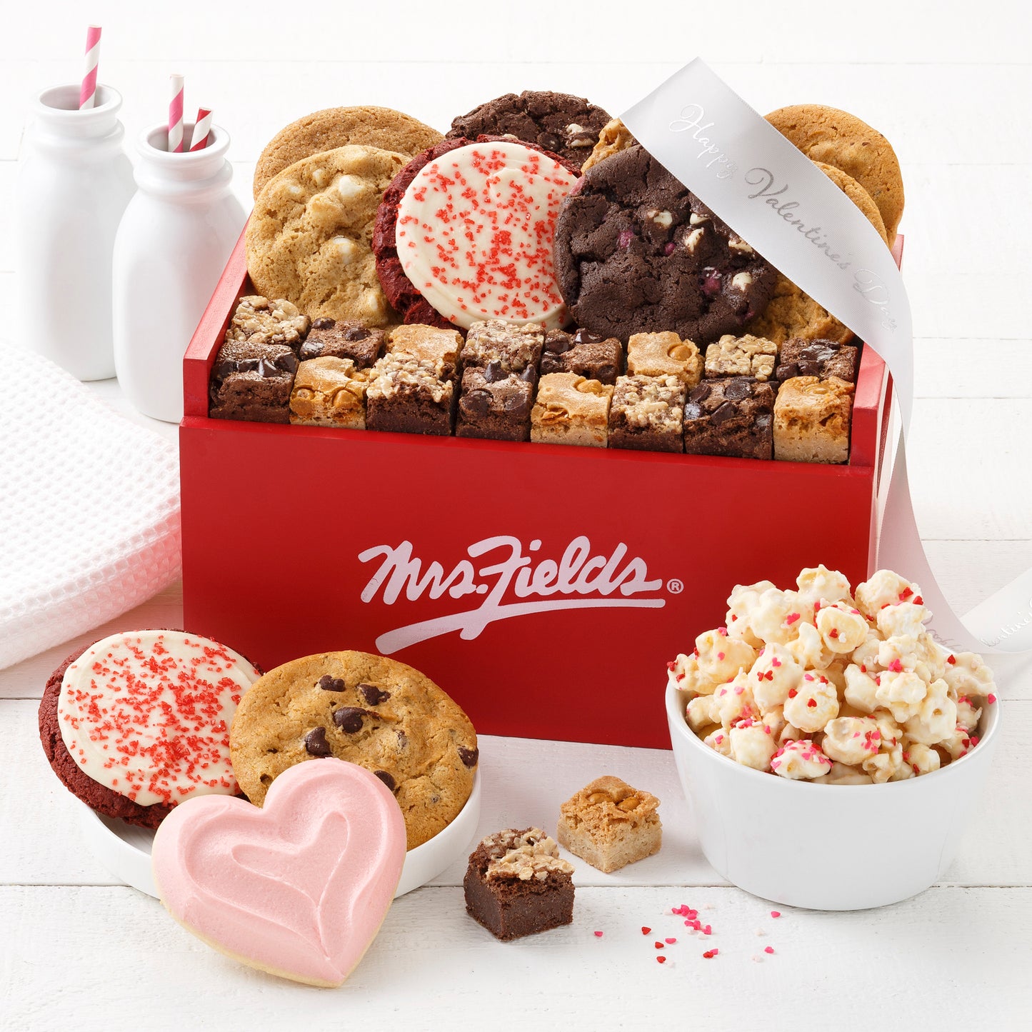 A red Mrs. Fields Signature wooden crate and filled with an assortment of original cookies, brownie bites, frosted cookies, and popcorn.