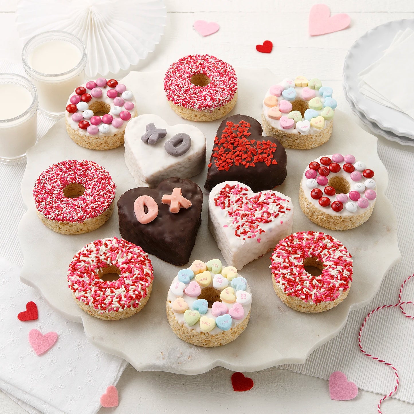 An assortment of jumbo Valentine's Day themed rice krispie treats each uniquely decorated, including four heart shaped, chocolate-covered rice krispies and eight donut shaped rice krispies decorated with sprinkles and candy.