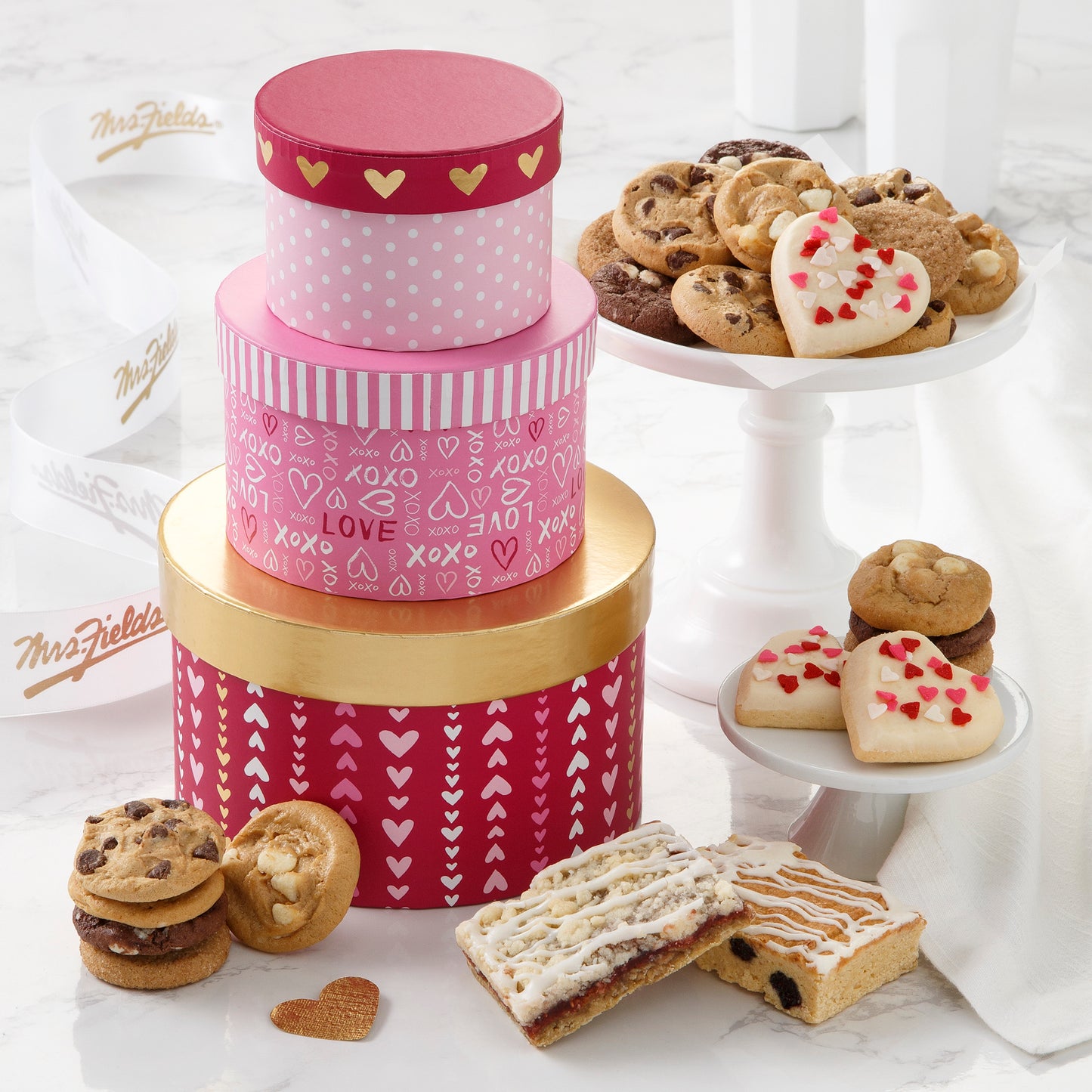 A pink, red, and gold three tier mini tower decorated with hearts and love sentiments and surrounded with nibblers, mini frosted cookies, and fruit bars.