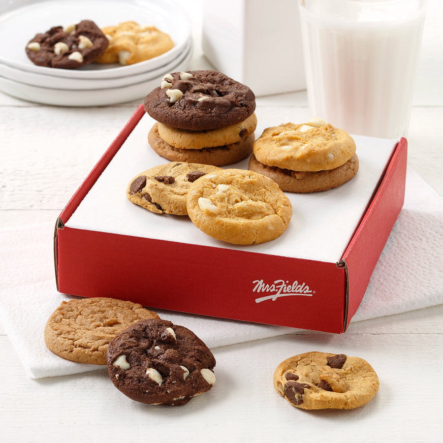 Red and white cardboard box with the Mrs. Fields logo on the side surrounded by an assortment of NIbblers bite-sized cookies with two glasses of milk on a white surface.