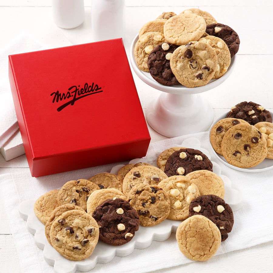 A square red gift box with a shiny Mrs. Fields logo on it surrounded by an assortment of small cookies on a white wooden surface with white serving trays and plates.