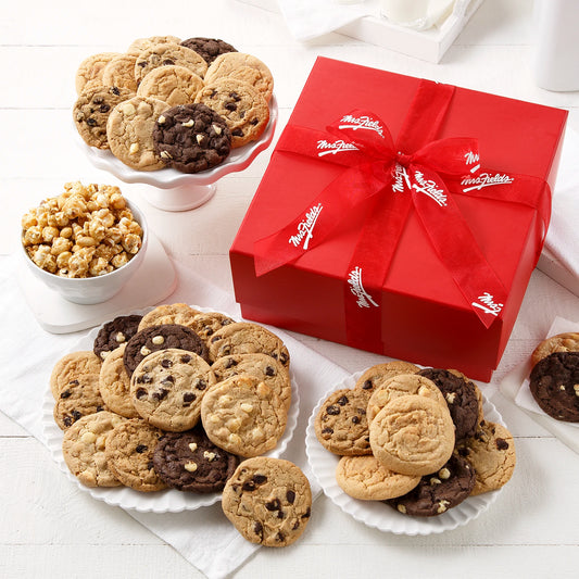 Red gift box tied with a Mrs. Fields red ribbon surrounded by an assortment of 38 Mrs. Fields cookies and toffee nut popcorn on a white surface with white dishes.