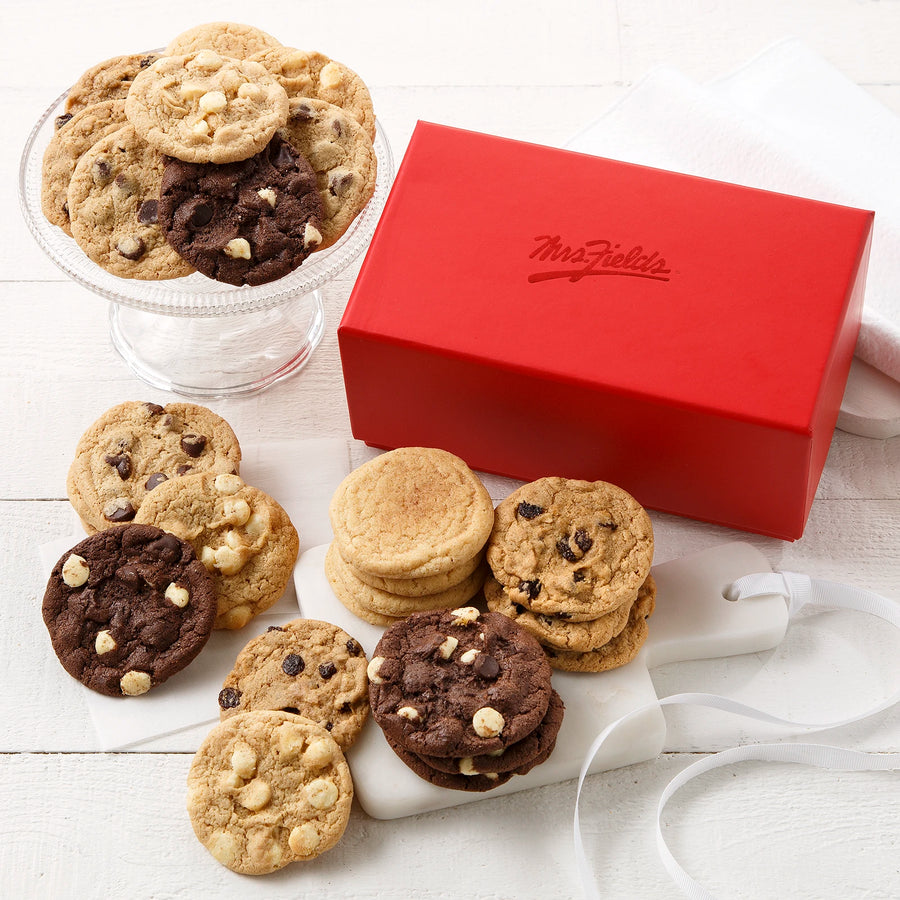 A rectangular red gift box with a shiny Mrs. Fields logo on it surrounded by an assortment of Nibblers bite-sized cookies on a white wooden surface.