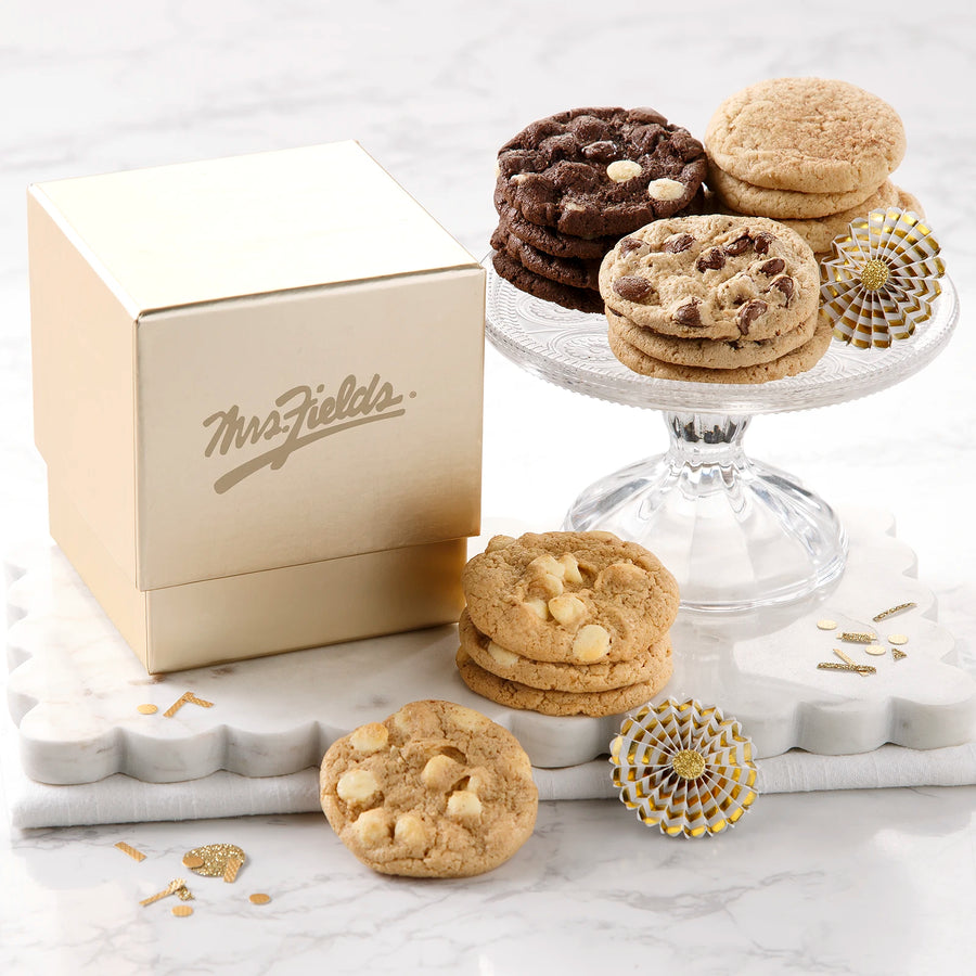 A square gold gift box with a shiny Mrs. Fields logo on it surrounded by an assortment of small cookies on a white marble surface with gold confetti.