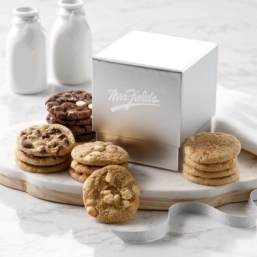A silver square gift box with a shiny Mrs. Fields logo on it surrounded by an assortment of Nibblers bite-sized cookies on a white marble surface.
