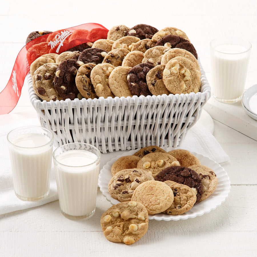 A white basket full of Nibblers bite-sized cookies with a red Mrs. Fields ribbon and two glasses of milk on a white wooden surface.,