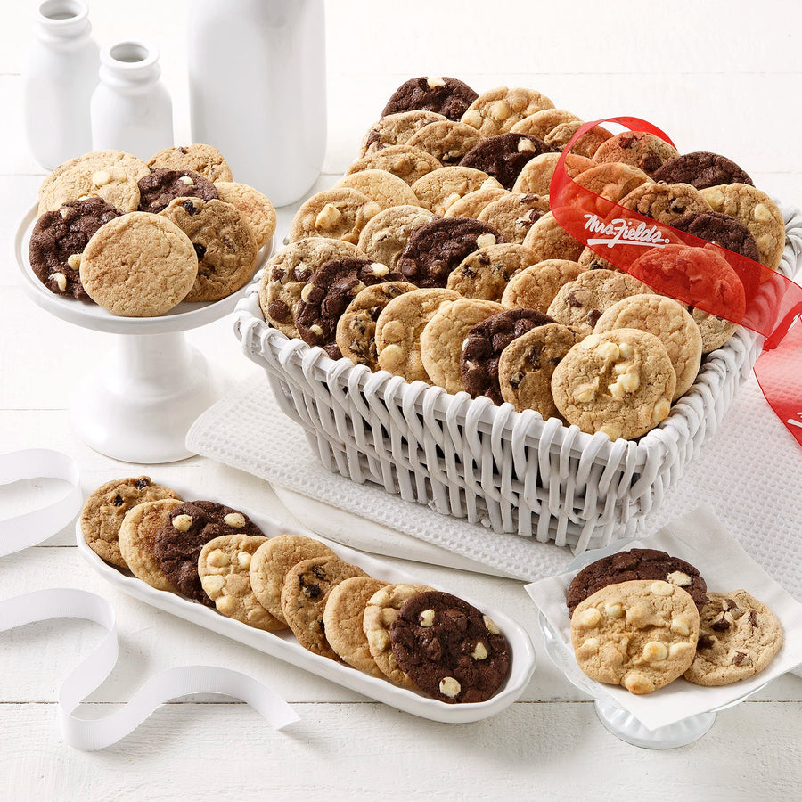 A white basket filled with an assortment of Nibblers bite-sized cookies with a red Mrs. Fields ribbon on a white wooden surface with white plates, napkins, and milk bottles.
