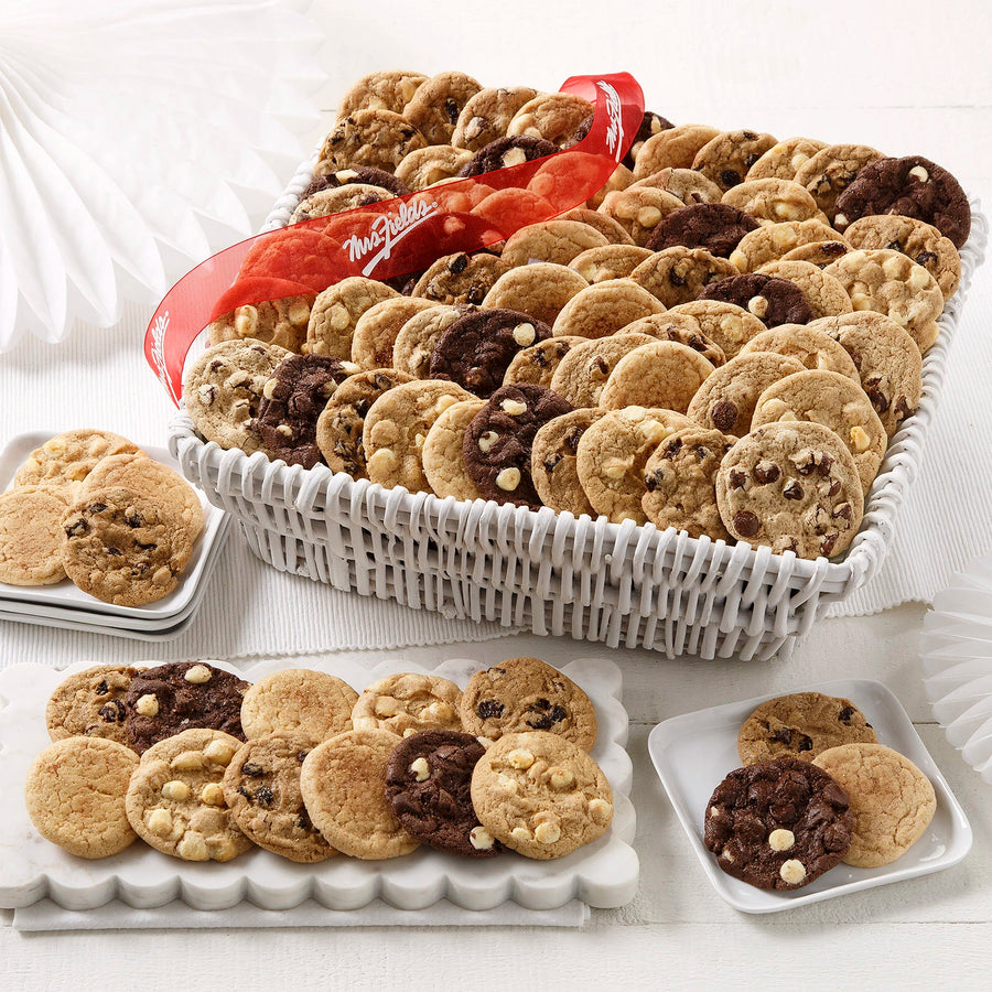 A large white wicker basket filled with an assortment of Nibblers bite-sized cookies on a white wooden surface with white plates and a red Mrs. Fields ribbon.
