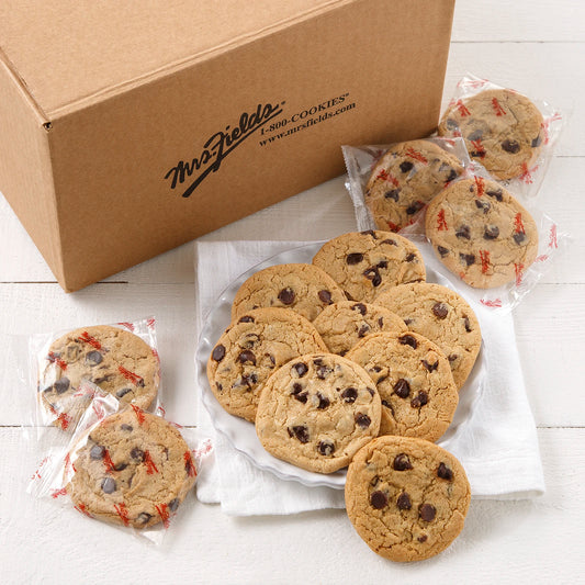 A large mailer box of Mrs. Field's chocolate chip cookies with several cookies displayed on a white plate and wrapped individually in Mrs. Fields printed wrapping on a white surface.