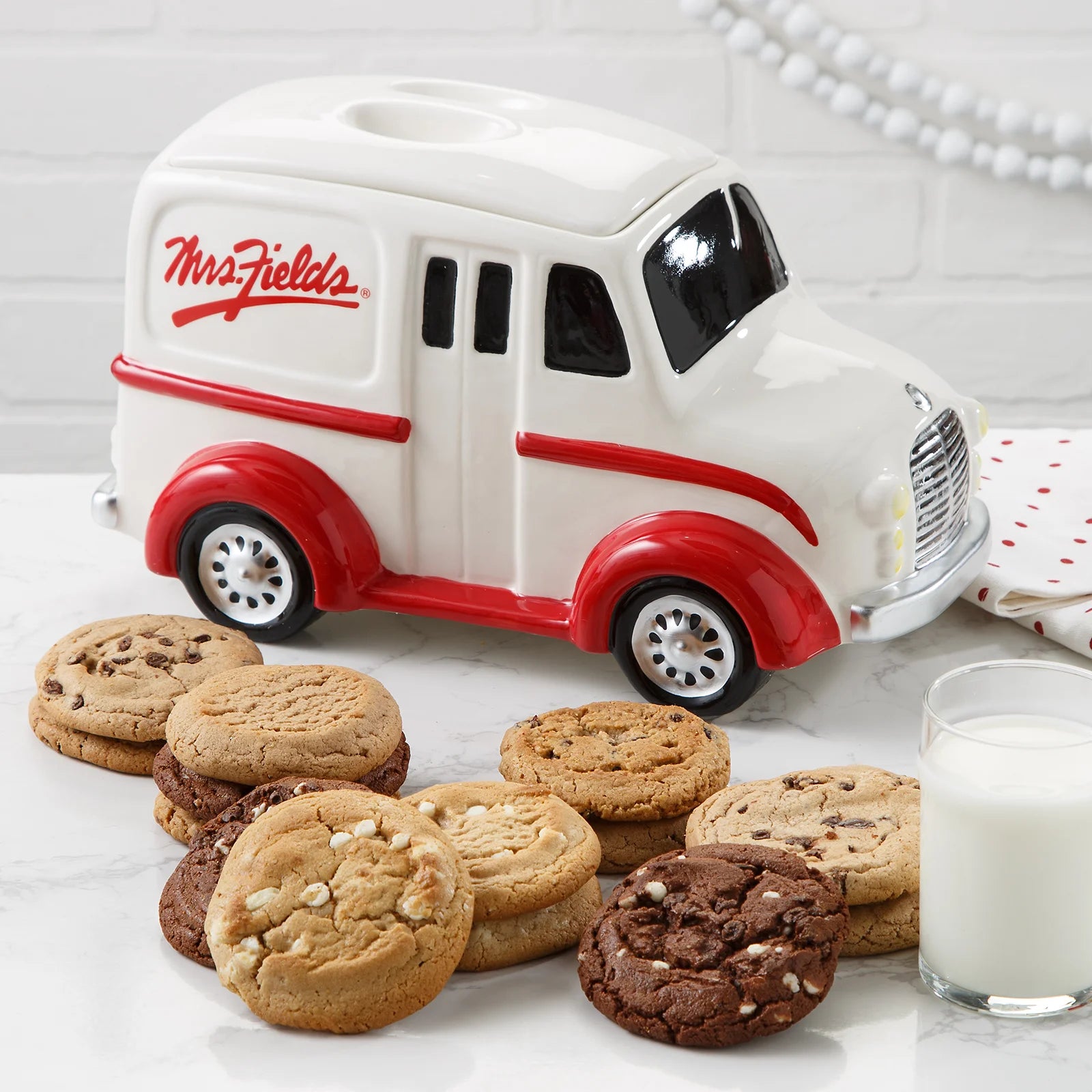 Red and white Mrs. Fields branded novelty cookie jar in the shape of a milk truck surrounded by an assortment of cookies in five flavors including chocolate chip and triple chocolate on a white surface with a glass of milk.