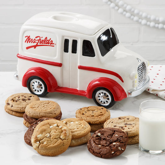 Red and white Mrs. Fields branded novelty cookie jar in the shape of a milk truck surrounded by an assortment of cookies in five flavors including chocolate chip and triple chocolate on a white surface with a glass of milk.