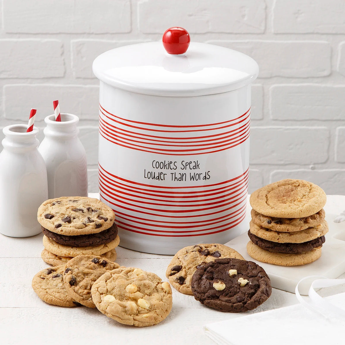 Ceramic cylindrical white and red cookie jar that says "cookies speak louder than words" on it surrounded by an assortment of fourteen Mrs. Fields original cookies.