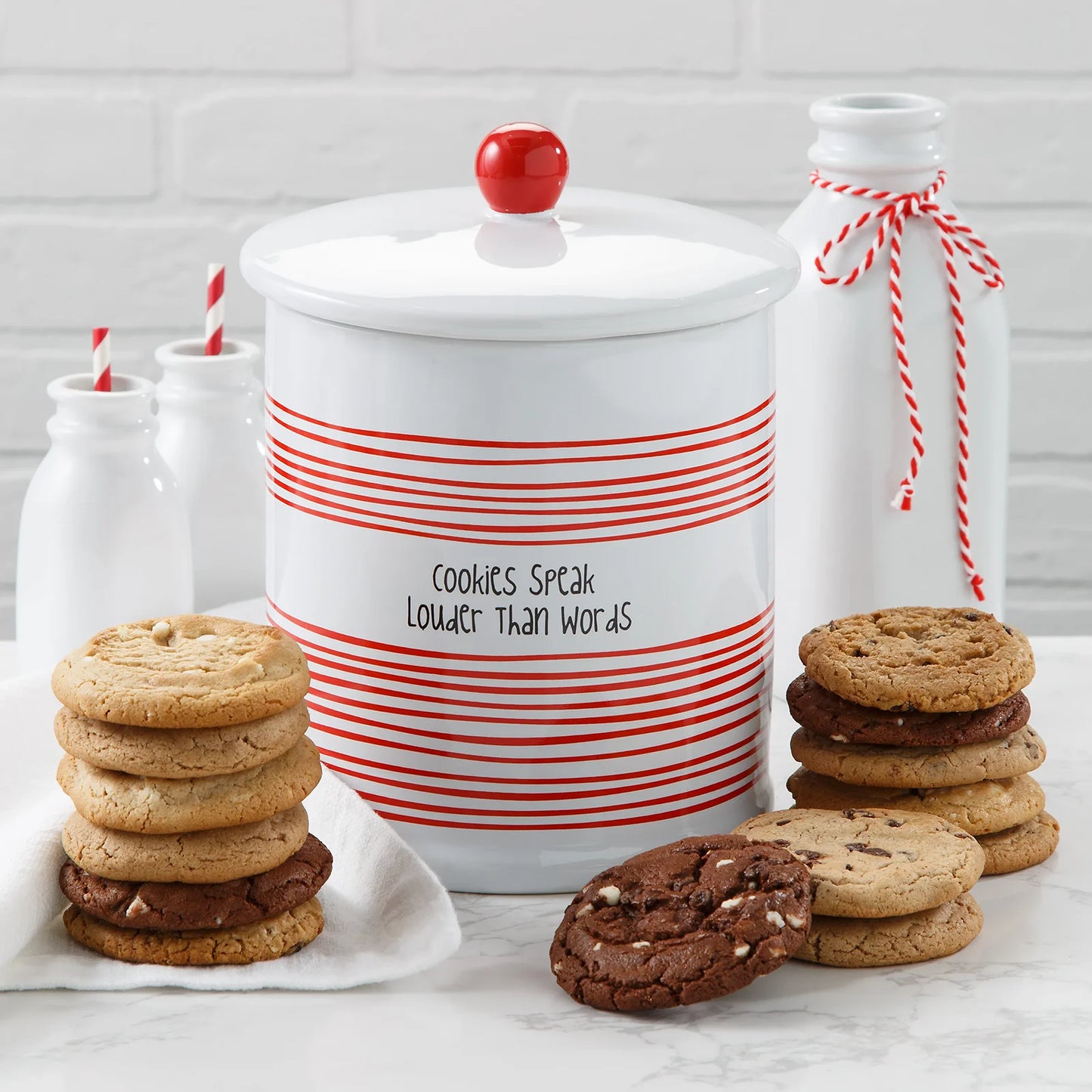A white and red cookie jar that says "Cookies Speak Louder Than Words" on it surrounded by an assortment of Mrs. Fields cookies in five flavors including chocolate chip and triple chocolate on a white surface with white milk bottles.