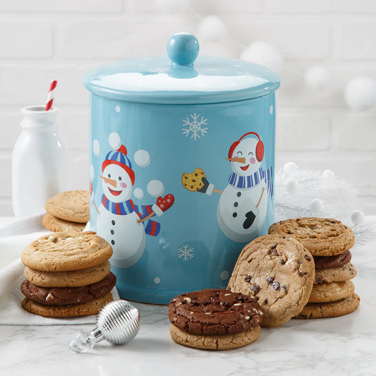 Blue holiday themed cookie jar with snowflakes and snowmen in winter hats and scarves on it surrounded by Mrs. Fields original cookies in five flavors including chocolate chip and triple chocolate on a white surface with holiday ornaments.