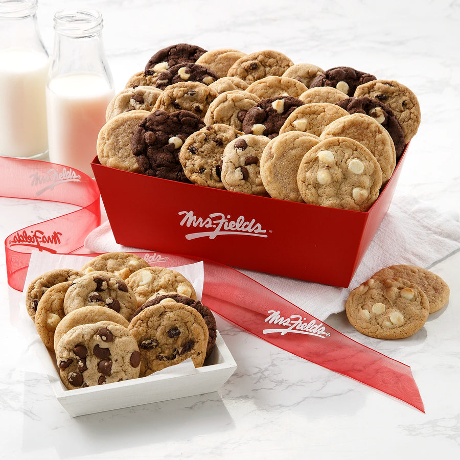 A red Mrs. Fields cookie crate filled with an assortment of Nibblers bite-sized cookies with a red Mrs. Fields ribbon and two bottles of milk on a white marbled surface.