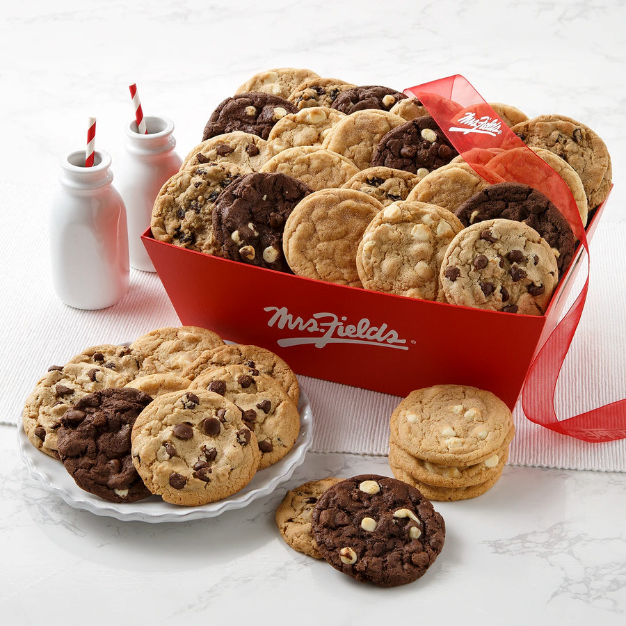 A red Mrs. Fields crate filled with an assortment of cookies with two white milk bottles and white plates on a white marble surface.