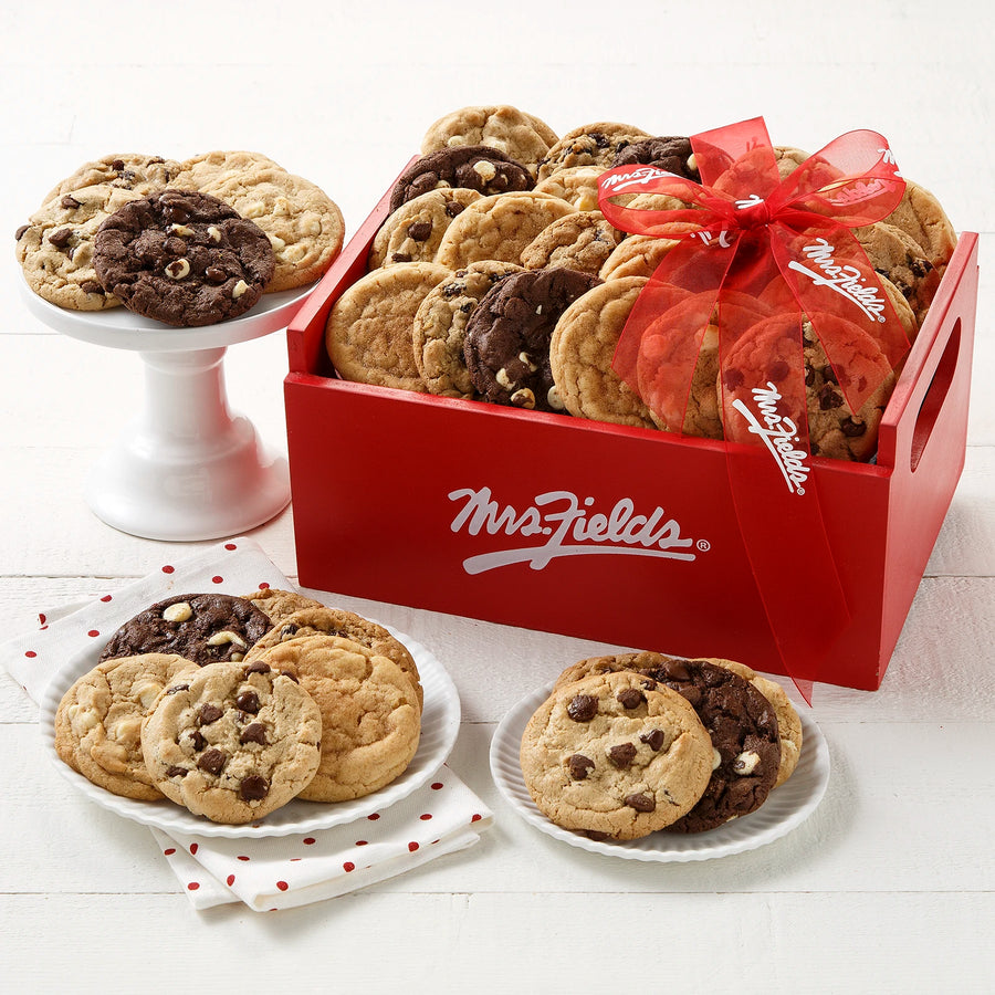 A red wooden Mrs. Fields crate filled with an assortment of cookies on a white wooden surface with a red Mrs. Fields ribbon, white plates, and white and red polka dotted napkins.