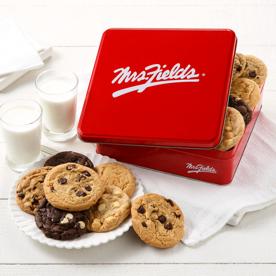 A red Mrs. Fields cookie tin with an assortment of a dozen cookies on a white surface with white plates, napkins, and two glasses of milk.