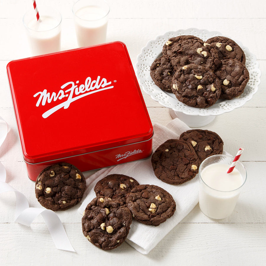 Mrs. Fields signature red cookie tin with a dozen triple chocolate cookies on a white surface with glasses of milk.