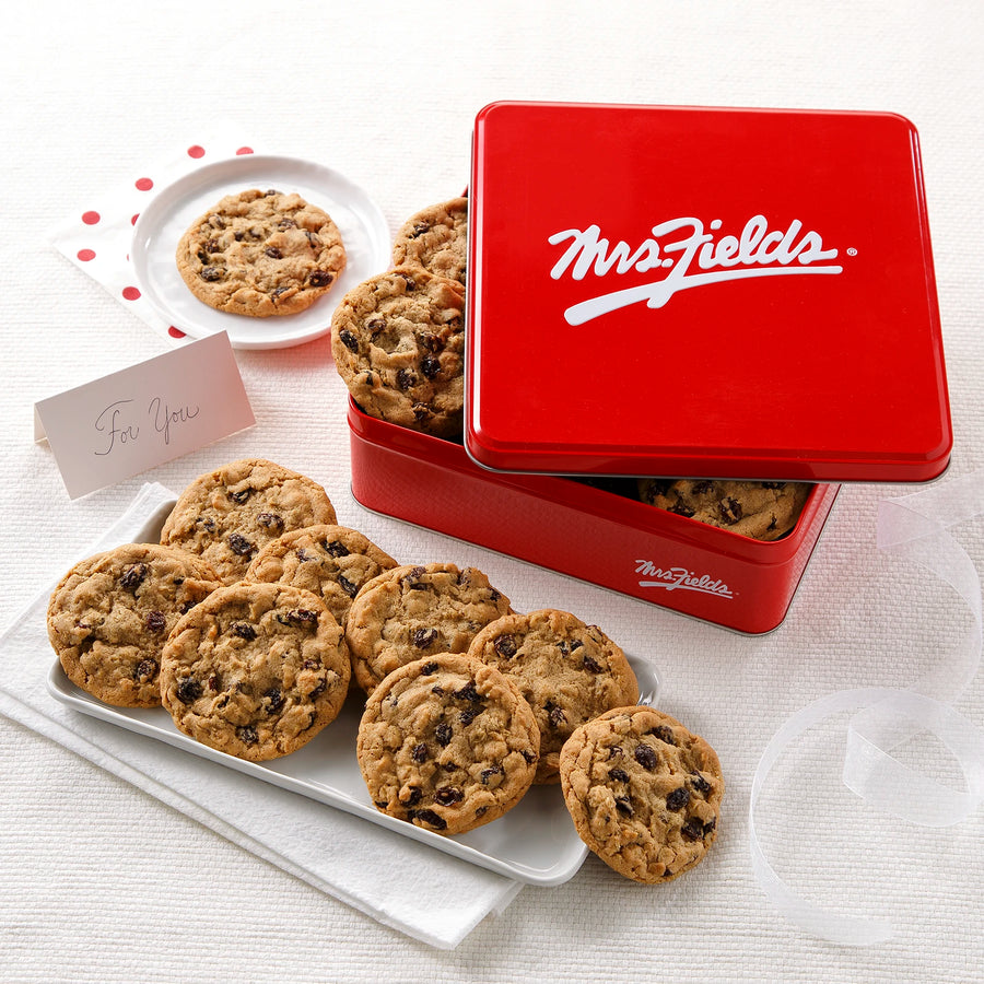 A red Mrs. Fields cookie tin with a dozen oatmeal raisin cookies on a white surface with white plates, napkins, and a small card that says "for you."