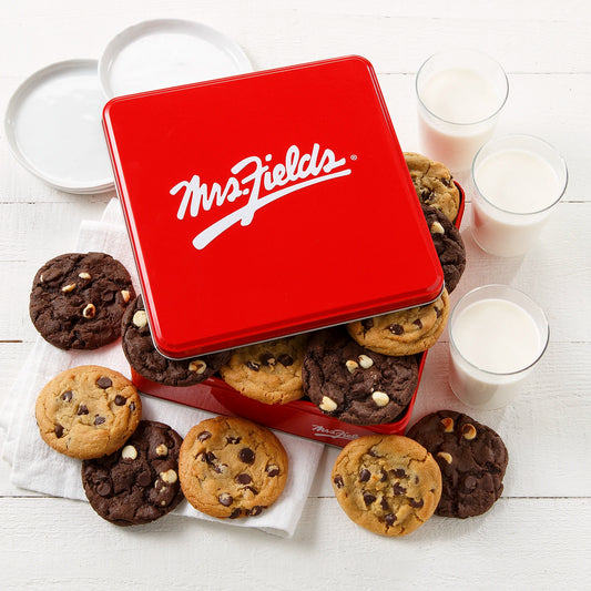 Mrs. Fields signature red cookie tin with a dozen semi-sweet chocolate chip and triple chocolate cookies on a white surface with plates and glasses of milk.