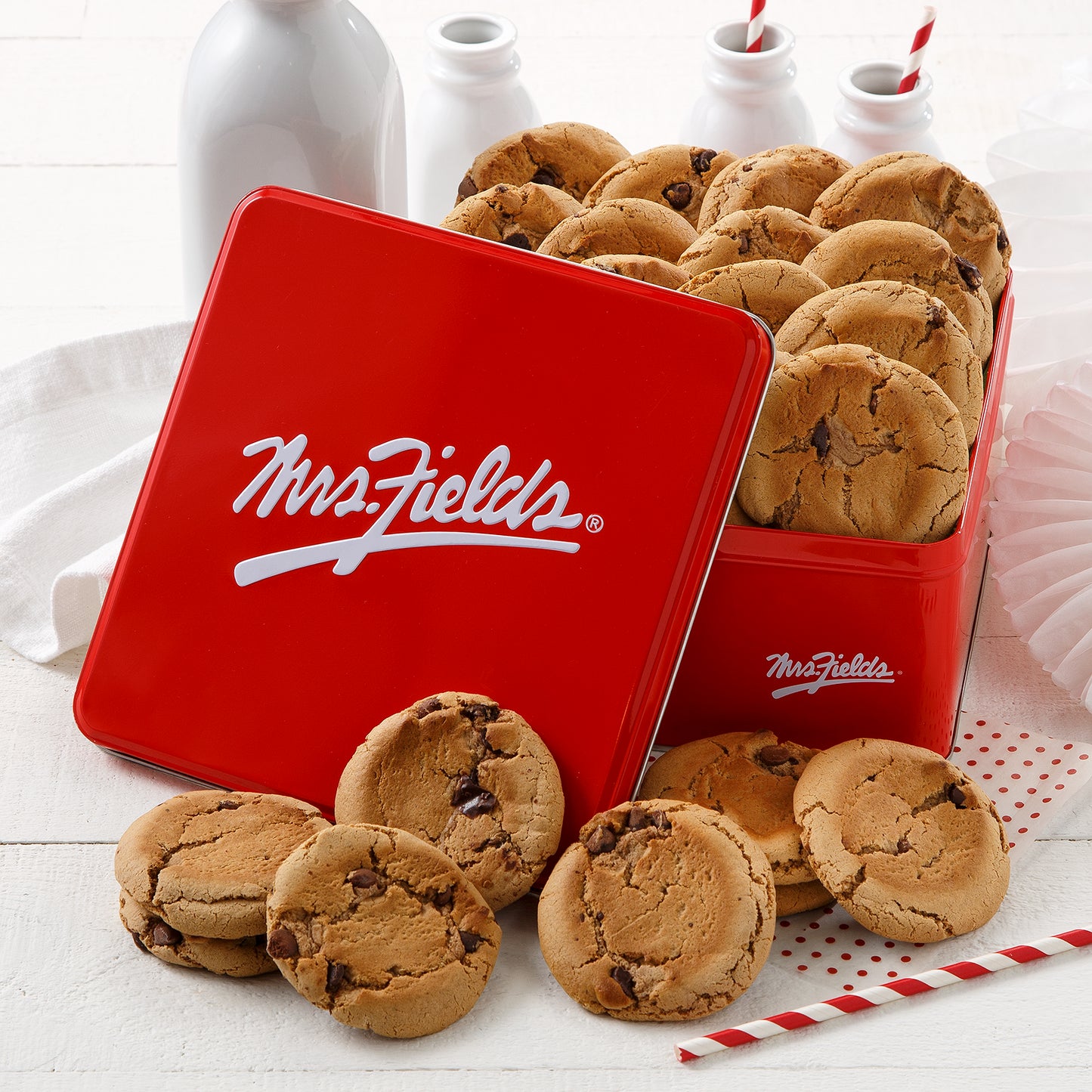 Mrs. Fields signature red tin with two dozen original chocolate chip cookies on a white surface with milk bottles.