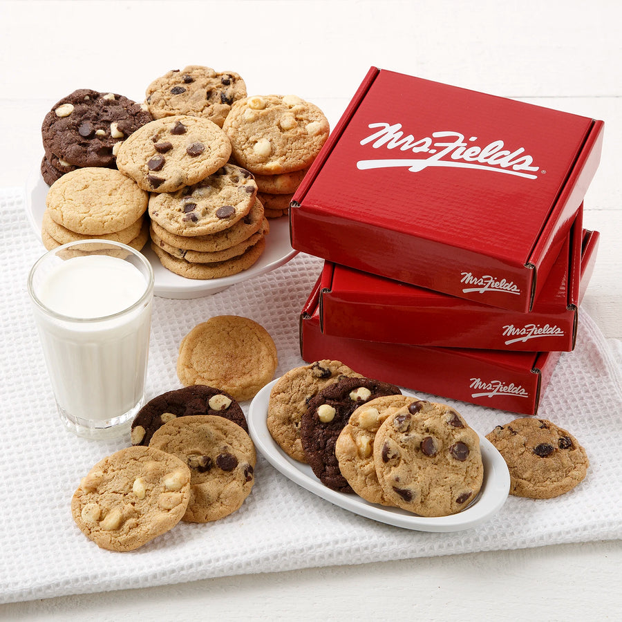 Three red boxes with a white Mrs. Fields logo on them stacked and surrounded by an assortment of small cookies on a white wooden surface.