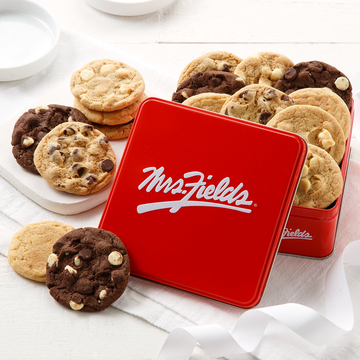 A red metal cookie tin with the white script logo 'Mrs. Fields' sits on a white surface, surrounded by assorted chocolate chip cookies.