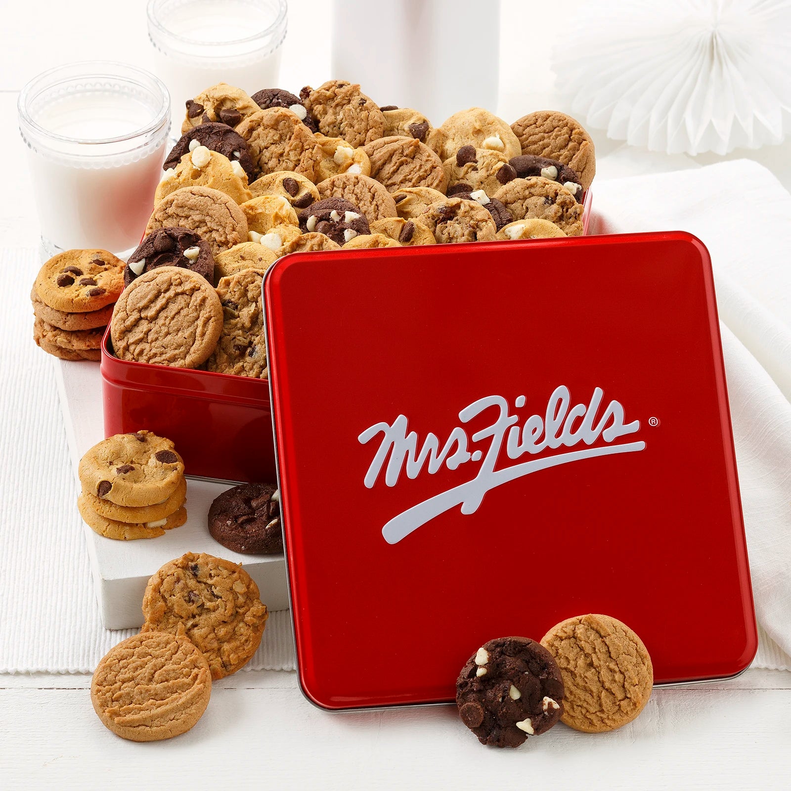 Mrs. Fields signature red cookie tin filled with an assortment of NIbblers bite-sized cookies on a white surface with two glasses of milk.
