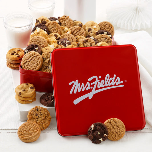 Mrs. Fields signature red cookie tin filled with an assortment of NIbblers bite-sized cookies on a white surface with two glasses of milk.