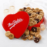 Mrs. Fields red heart-shaped cookie tin filled with an assortment of Nibblers bite-sized cookies in five flavors on a white surface with two champagne glasses.