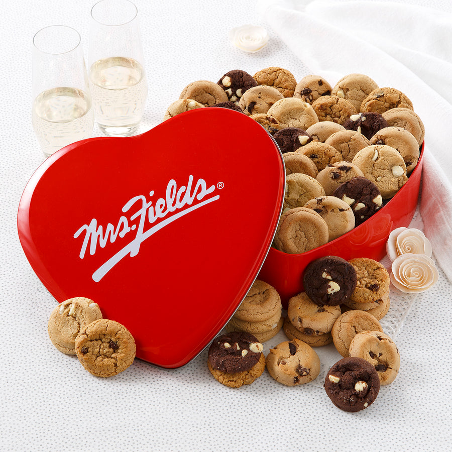 Mrs. Fields red heart-shaped cookie tin filled with an assortment of Nibblers bite-sized cookies in five flavors on a white surface with two champagne glasses.