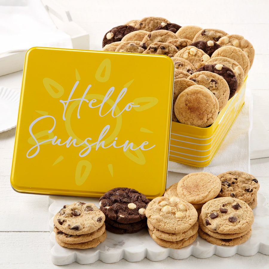 A yellow cookie tin that says "Hello Sunshine" on it in white cursive text is filled with an assortment of Nibblers bite-sized cookies on a white surface with a white marble scalloped serving platter.