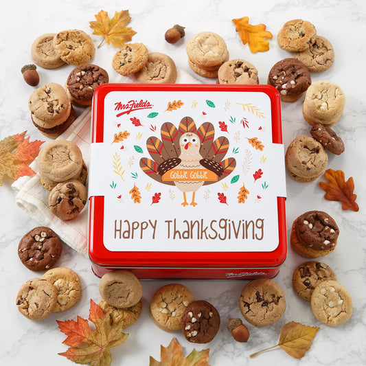 Cookie tin with a "Happy Thanksgiving" sleeve on it that has a cute cartoon turkey that says "gobble gobble" on it surrounded by Mrs. Fields Nibblers bite-sized cookies in four flavors including chocolate chip and triple chocolate on a white background with autumn leaves.