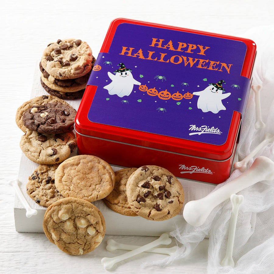 A red Mrs. Fields cookie tin with a purple Halloween-themed sleeve on it that says "Happy Halloween" with two ghosts holding a banner of jack-o-lanterns all next to an assortment of a dozen cookies on a white wooden surface with fake cob webs and bones.