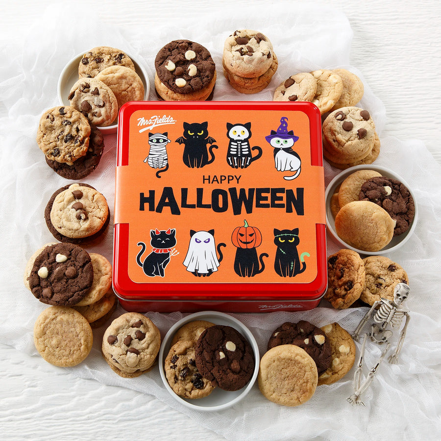 A red Mrs. Fields cookie tin with an orange Halloween-themed sleeve on it that says "Happy Halloween" with eight black cats in various costumes including a mummy, a bat, a witch and a ghost, all surrounded by an assortment of small cookies on a white surface with fake cob webs.