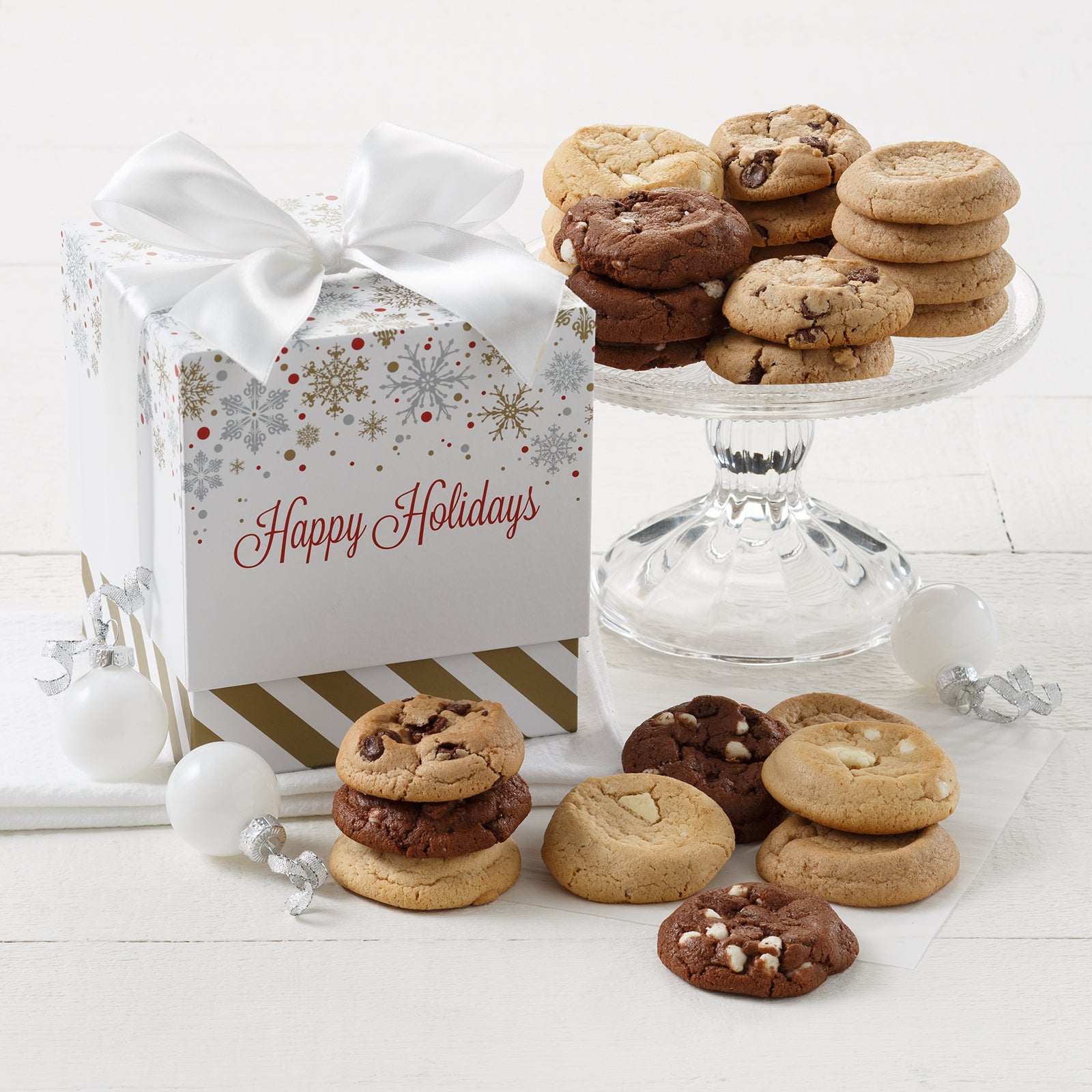 Mini cookie box that says "Happy Holidays" with a snowflake print on it surrounded by Mrs. Fields Nibblers bite-sized cookies in four flavors including chocolate chip and triple chocolate on a white surface with white Christmas ornaments.