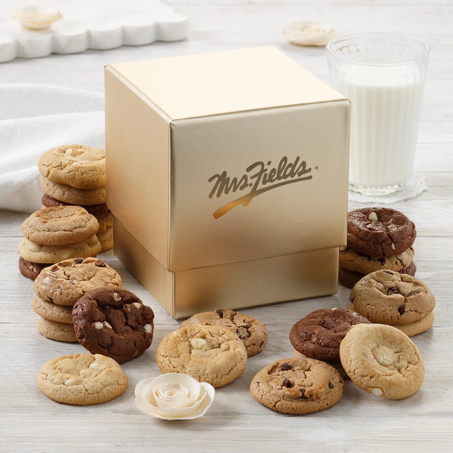Elegant gold Mrs. Fields mini cookie box surrounded by Nibblers bite-sized cookies in five flavors including chocolate chip and triple chocolate on a white surface with a glass of milk.