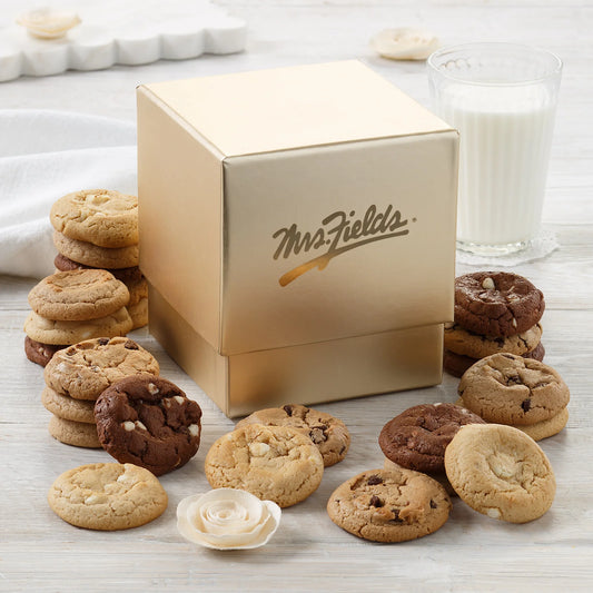 Elegant gold Mrs. Fields mini cookie box surrounded by Nibblers bite-sized cookies in five flavors including chocolate chip and triple chocolate on a white surface with a glass of milk.