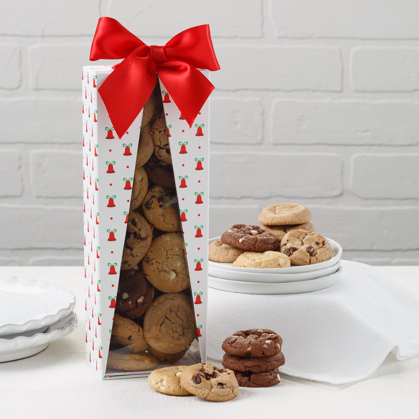An assortment of Mrs. Fields Nibblers bite-sized cookies in a holiday gift box with a red box on it and triangular cutout where the assortment of cookies inside are revealed. On a white surface with a stack of small white serving plates.
