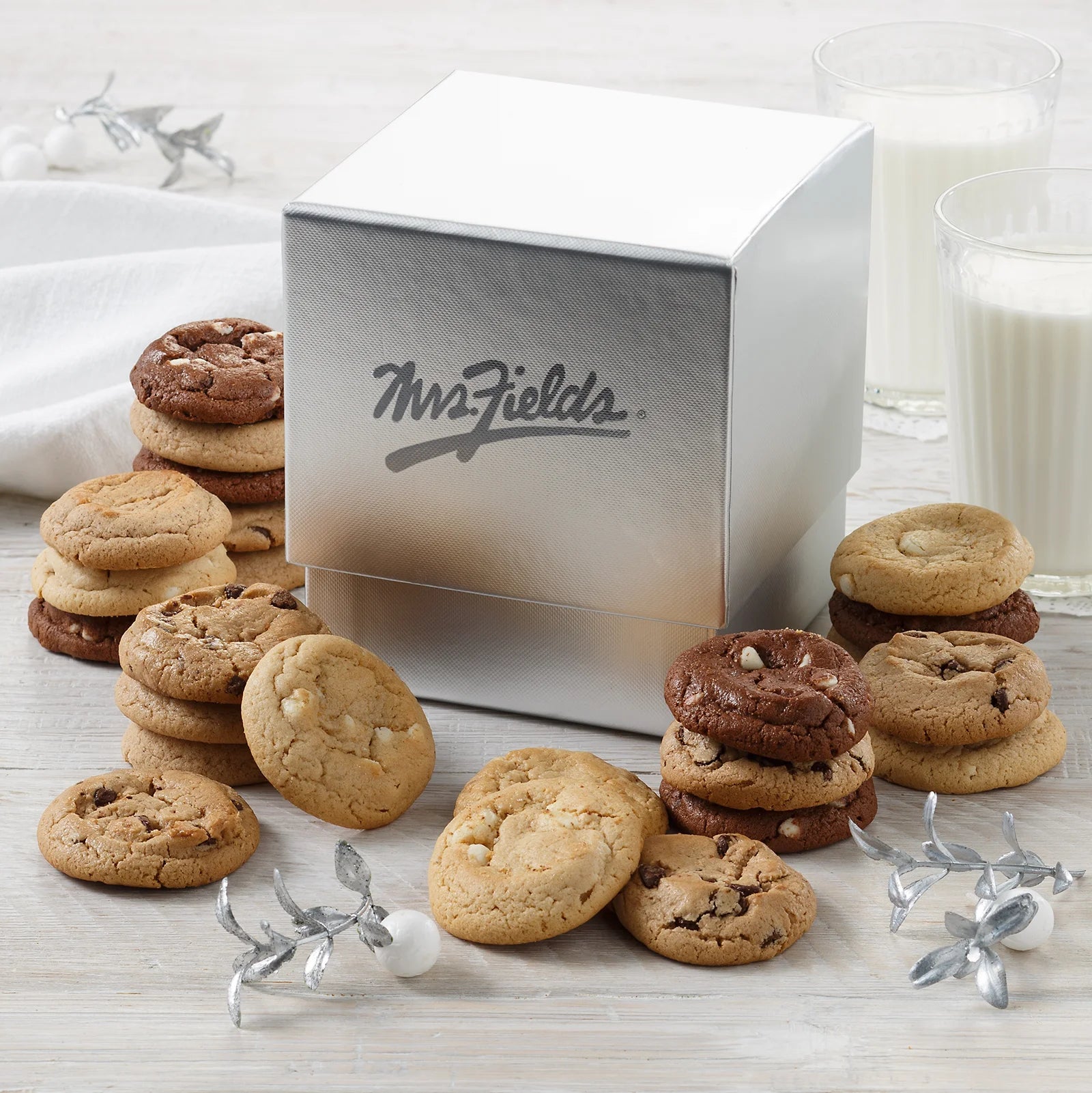 A shimmering silver Mrs. Fields mini cookie box surrounded by an assortment of Nibblers bite-sized cookies in five flavors including chocolate chip and white chocolate on a white surface with a glass of milk.