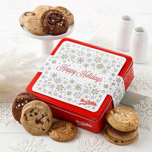 Mrs. Fields signature red cookie tin with a Happy Holidays sleeve on it surrounded by a dozen original cookies in five flavors including chocolate chip and white chocolate chip on a white surface with snowflake decorations and milk bottles.