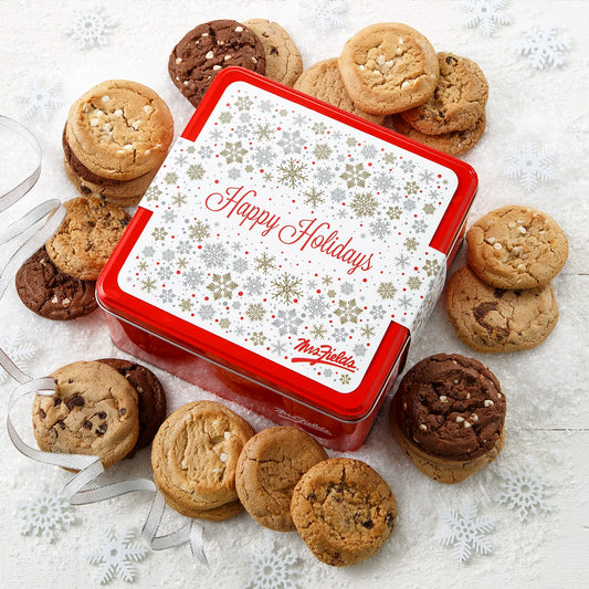Mrs. Fields signature red cookie tin with a Happy Holidays sleeve on it surrounded by two dozen original cookies in five flavors including chocolate chip and white chocolate chip on a white surface with snowflake decorations and silver ribbon.