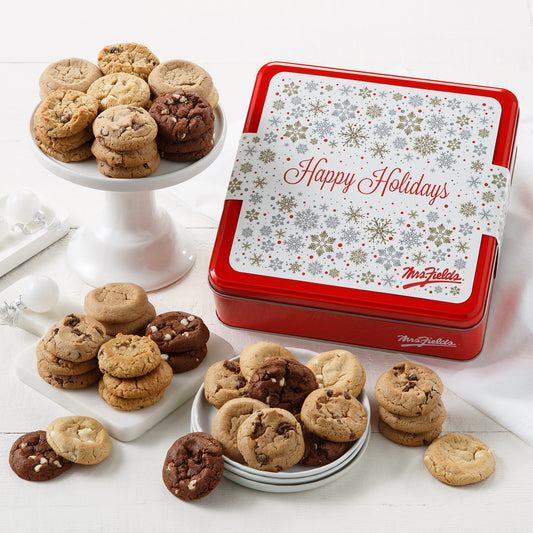 A Mrs. Fields signature red cookie tin with a snowflake themed sleeve that says "Happy Holidays" surrounded by an assortment of Nibblers bite-sized cookies in five flavors including chocolate chip and triple chocolate on a white surface with white Christmas ornaments.