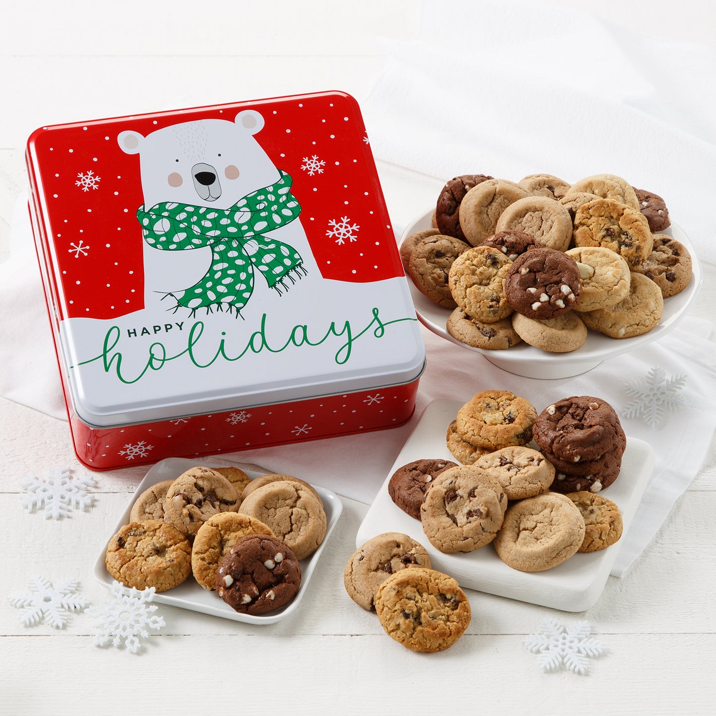 Red cookie tin that says "Happy Holidays" on it with an image of a polar bear in a scarf surrounded by an assortment of Mrs. Fields Nibblers bite-sized cookies in five flavors including chocolate chip and oatmeal raising on a white surface with snowflake cutouts.