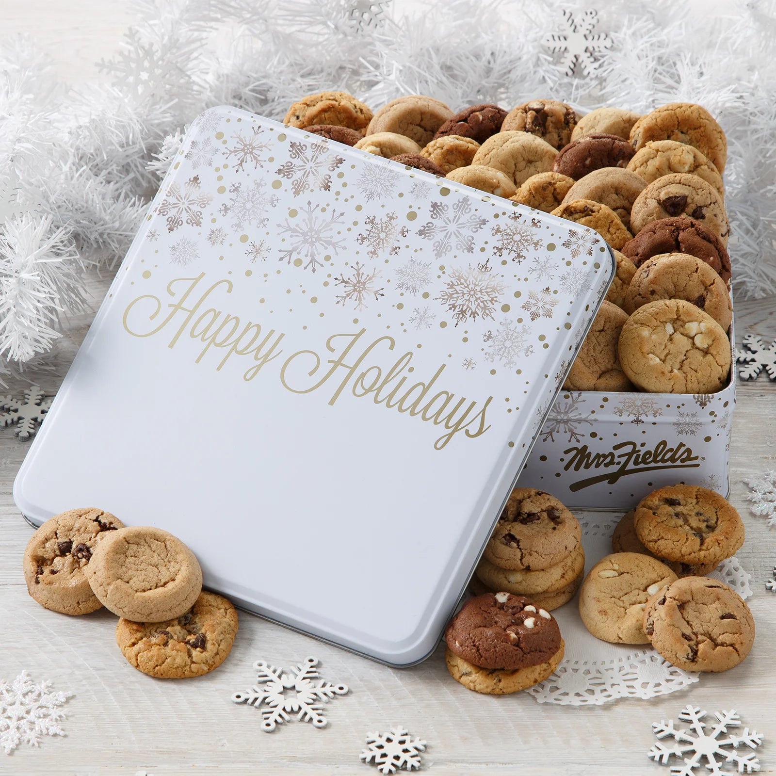 A "Happy Holidays" snowflake pattern cookie tin filled with Mrs. Fields Nibblers bite-sized cookies in five flavors including chocolate chip and triple chocolate on a white surface with snowflake cutouts 